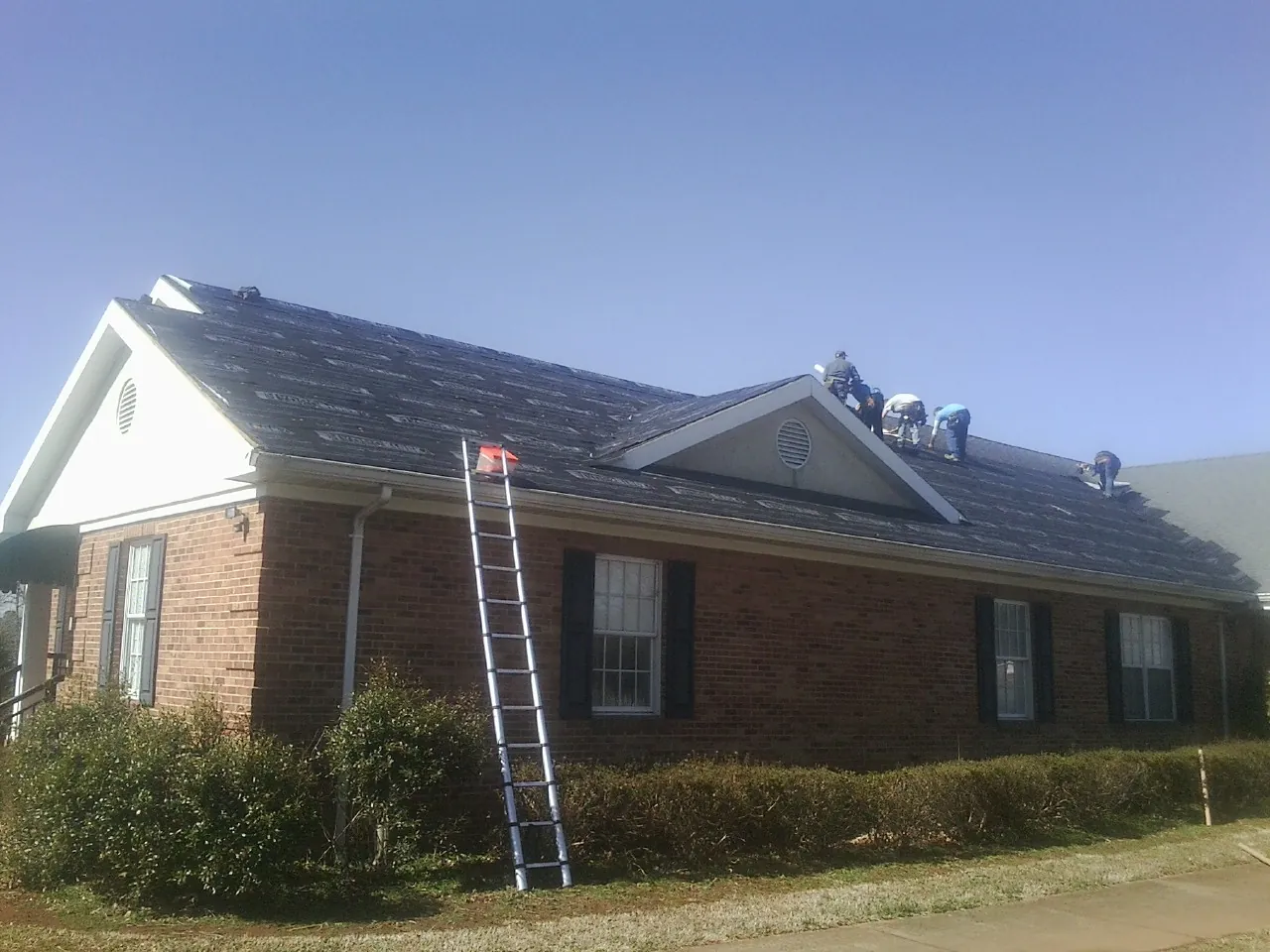 Crew tearing shingles off a brick ranch roof with a ladder set — Emergency Roof Repair work by our crew in Sauquoit