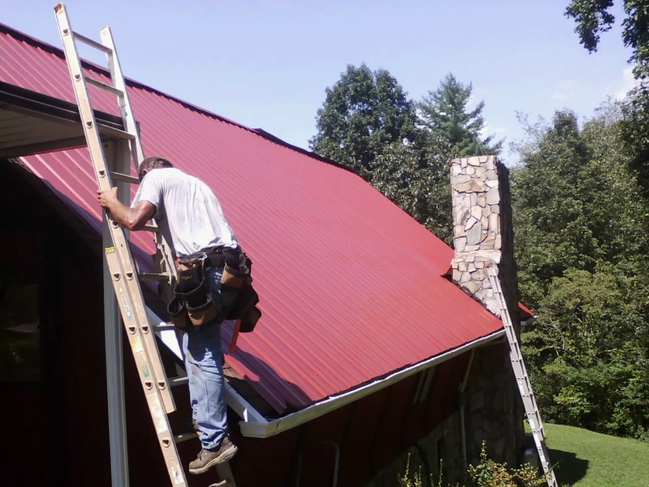 Roofer on a ladder beside a red metal roof and stone chimney — Soffit & Fascia Repair by Roofing Brothers in Sauquoit